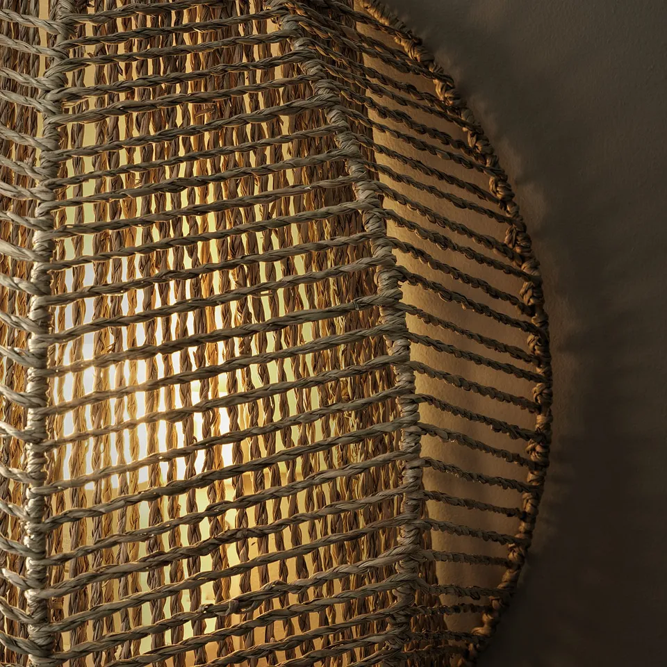 Hand-braided wall lamp, MÅNALG, emits warm light. Sedge material, brown-grey, hexagon shape, casts intricate shadows.