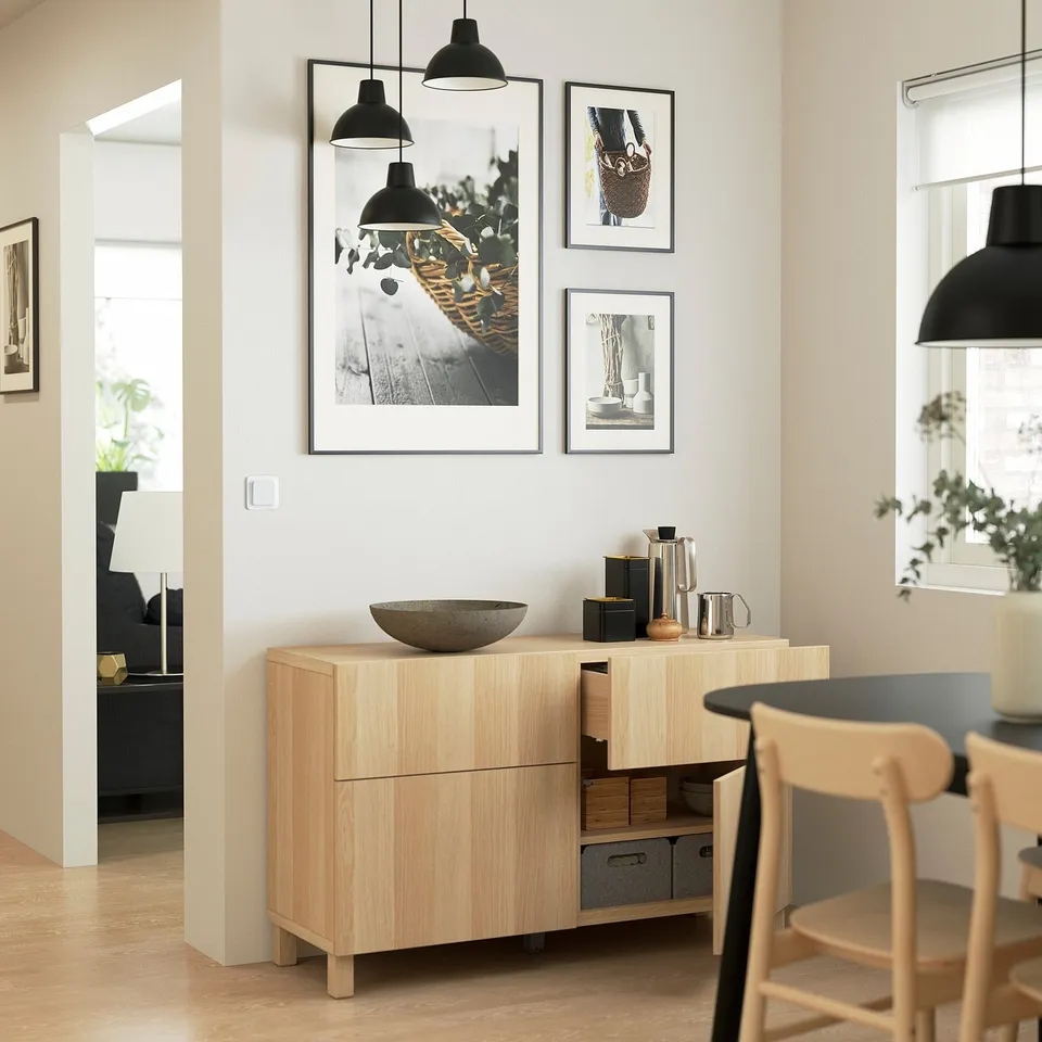Modern wooden sideboard with minimalist black pendant lights, three framed artworks, and a black kettle on top. Open drawer reveals shelves inside.