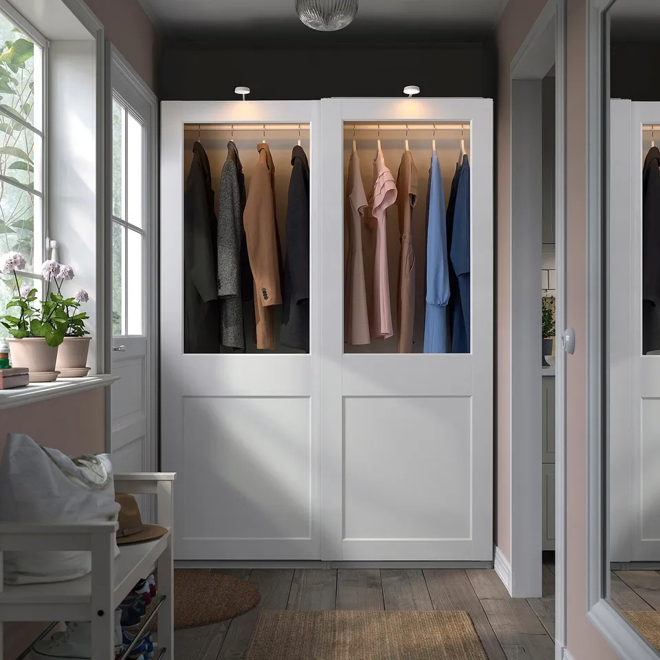 Modern PAX wardrobe with white sliding doors, showcasing clothes, taller than adjacent chair and closet, lit glass panels highlight contents.