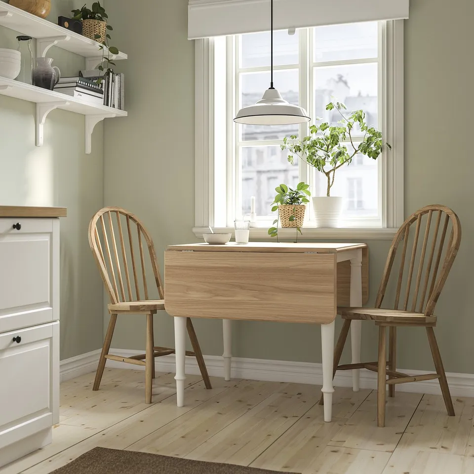 Wood drop-leaf table with white legs, chairs, books, and vases near a large window. Cosy and elegant for small spaces.