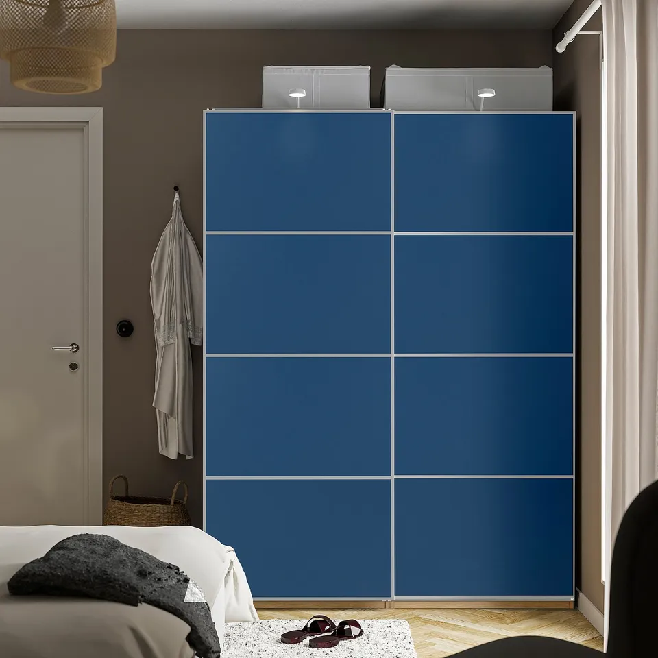 Modern bedroom with tall blue PAX wardrobe. Sliding doors, hanging clothes, and shoes on floor. Bed and door visible.