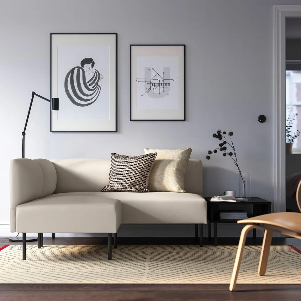 Modern LILLEHEM sofa in beige with 2 patterned pillows, black lamp, side table, and wall art. Minimalist room with grey walls, wooden chair, and rug.