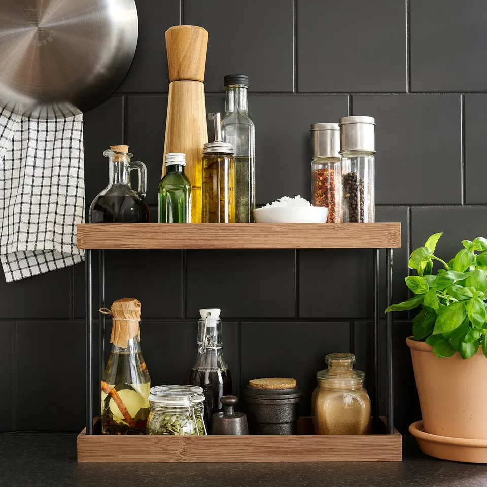 Wooden kitchen shelf holding spice jars, bottles, and a plant against black tiles.