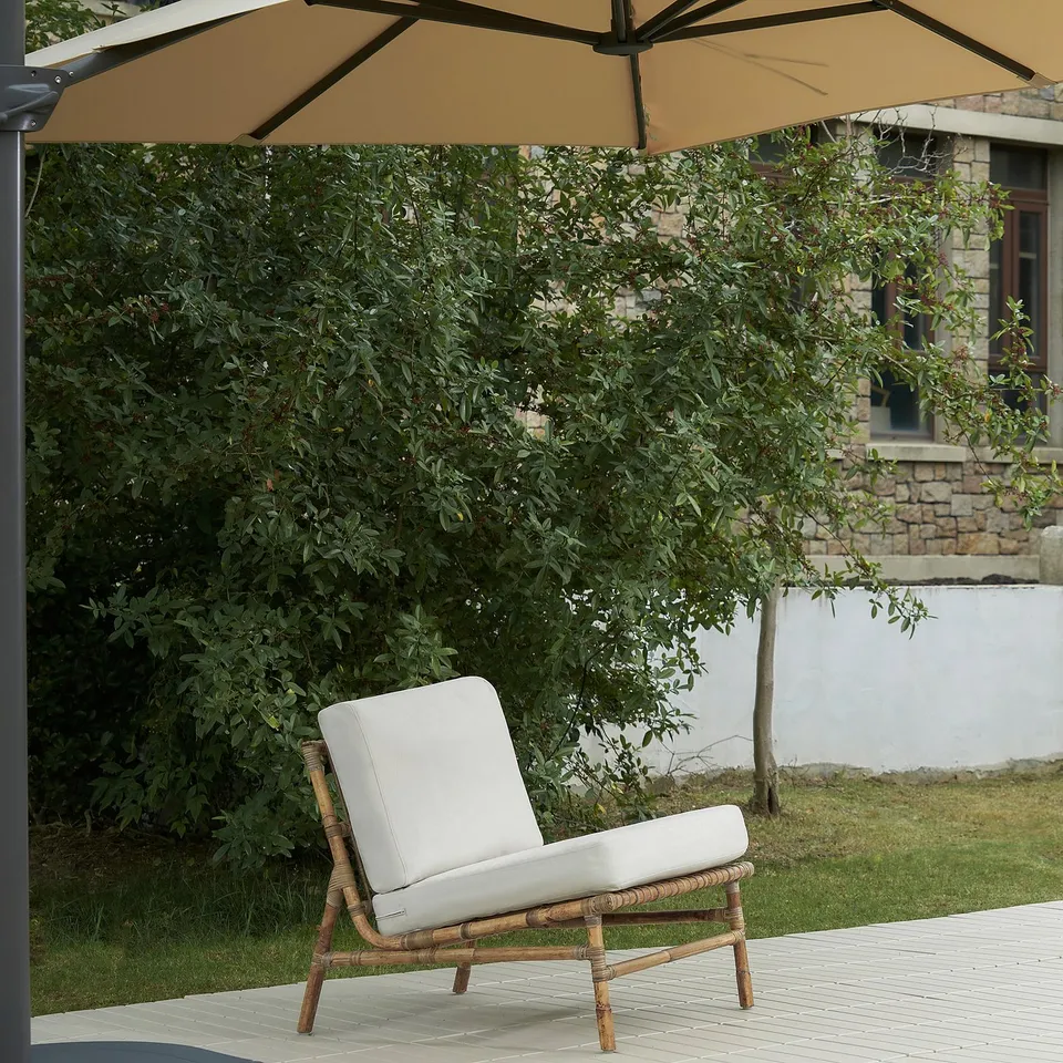Wicker chair with cushion under large umbrella on deck, beside greenery and stone building.
