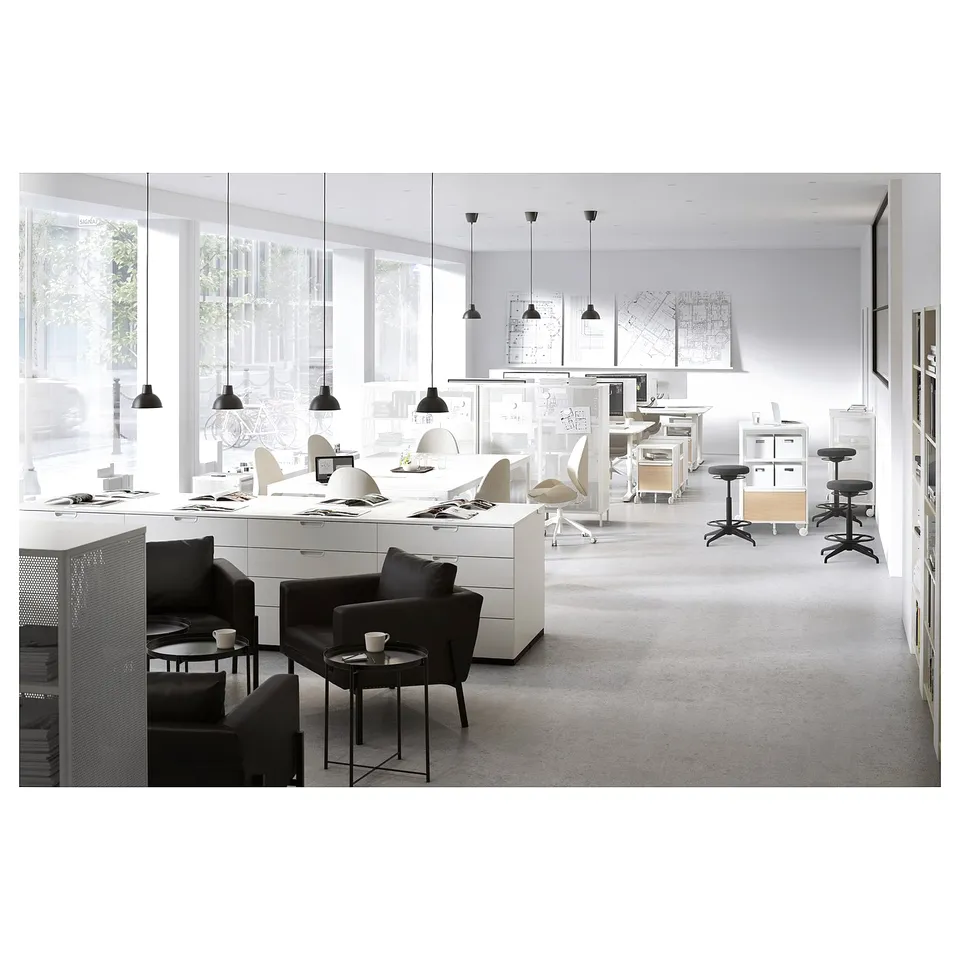 Modern office space with white desks, black chairs, and hanging lamps. Large windows provide natural light.