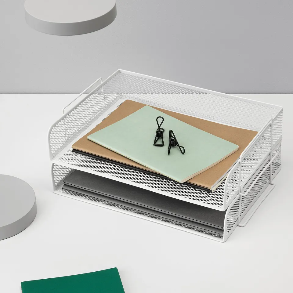 White metal mesh letter tray with two compartments, holding green folder, paper clips, and black binder clips, arranged neatly on a white desk.