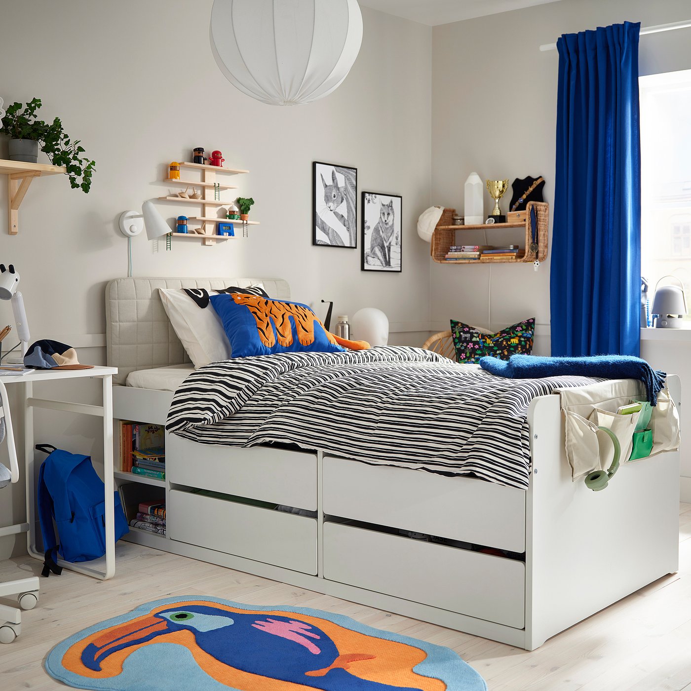 Childs room with toucan rug, blue & orange pillows, shelves with books, trophy, plant, blue curtains, desk, and chair.