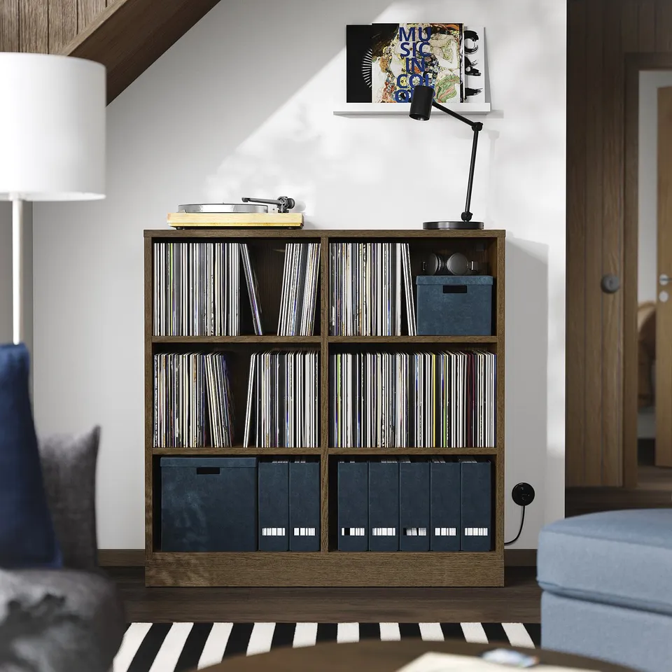 A TONSTAD wooden shelf unit with vinyl records and storage boxes. Its brown, rectangular, and sits between furniture, with a lamp and chair nearby.