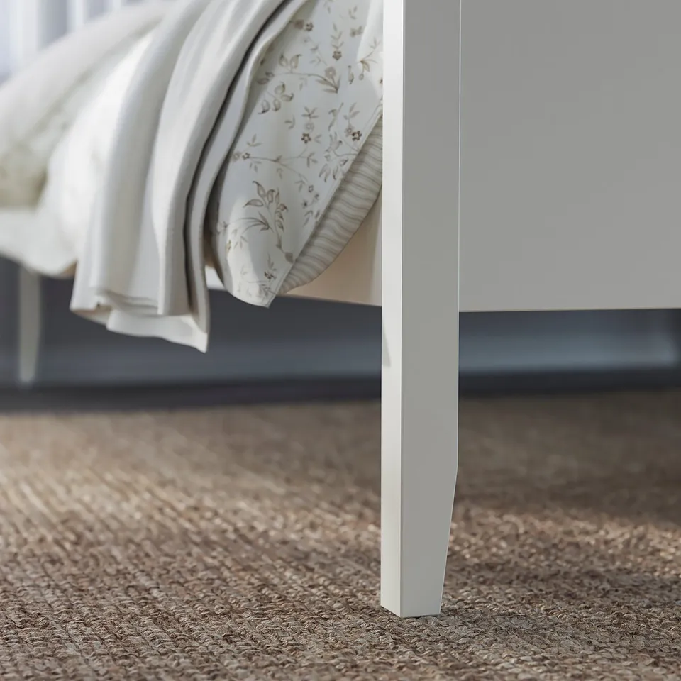 A white IDANÄS bed frame with a headboard, featuring slatted design. Light brown cushions sit against it, on a textured beige carpet.