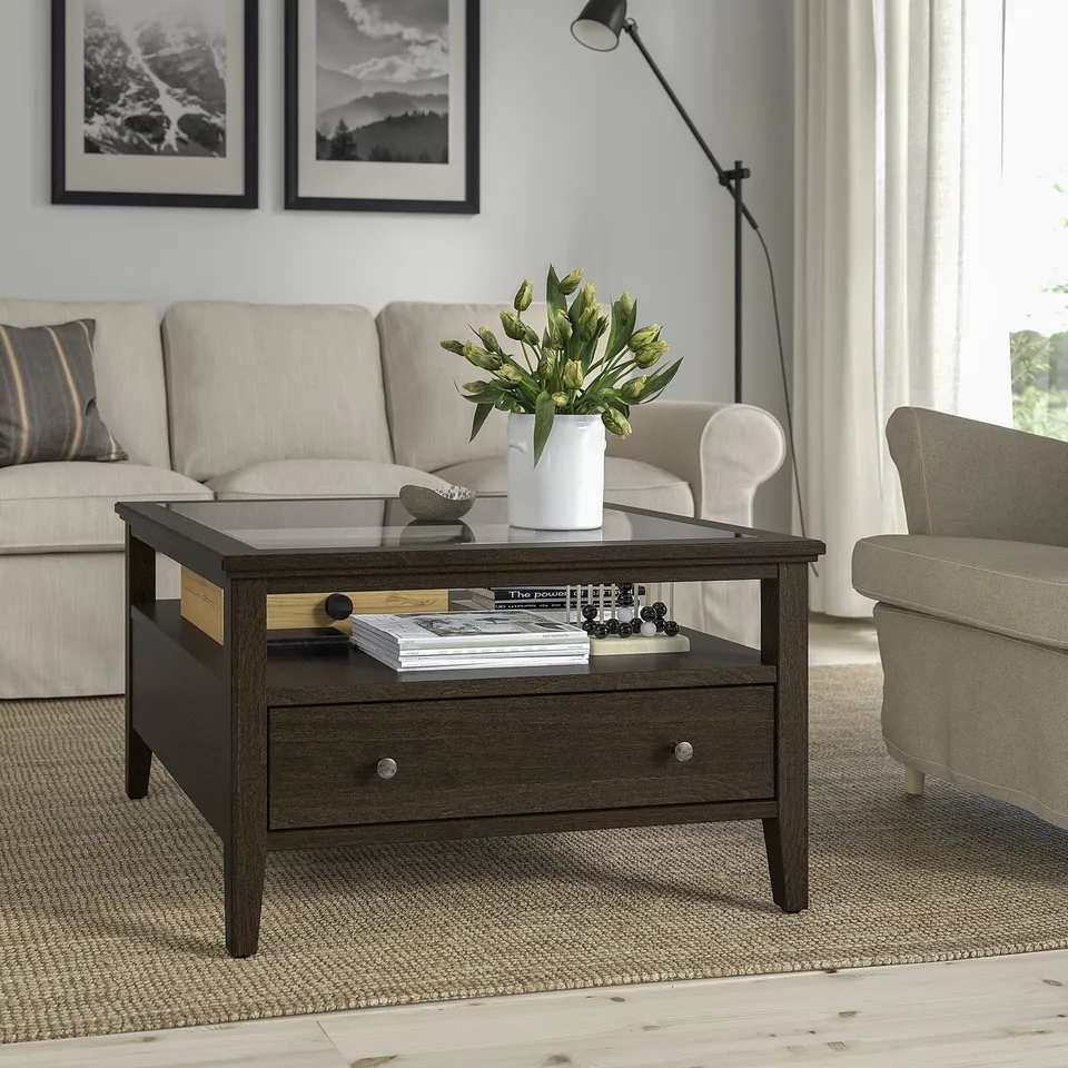 IDANÄS brown wood coffee table with glass top and drawer. Between beige sofa & armchair, holding books, vase & bowl. Moderate size.