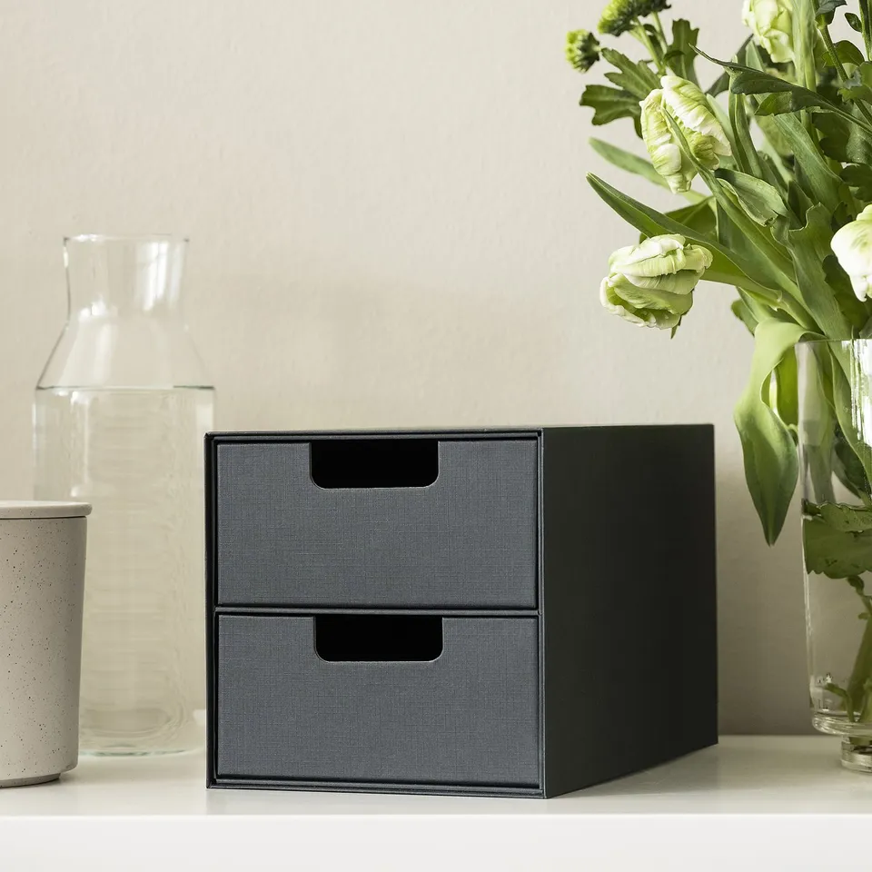 Black mini chest with two drawers on a white surface, surrounded by decorative items.