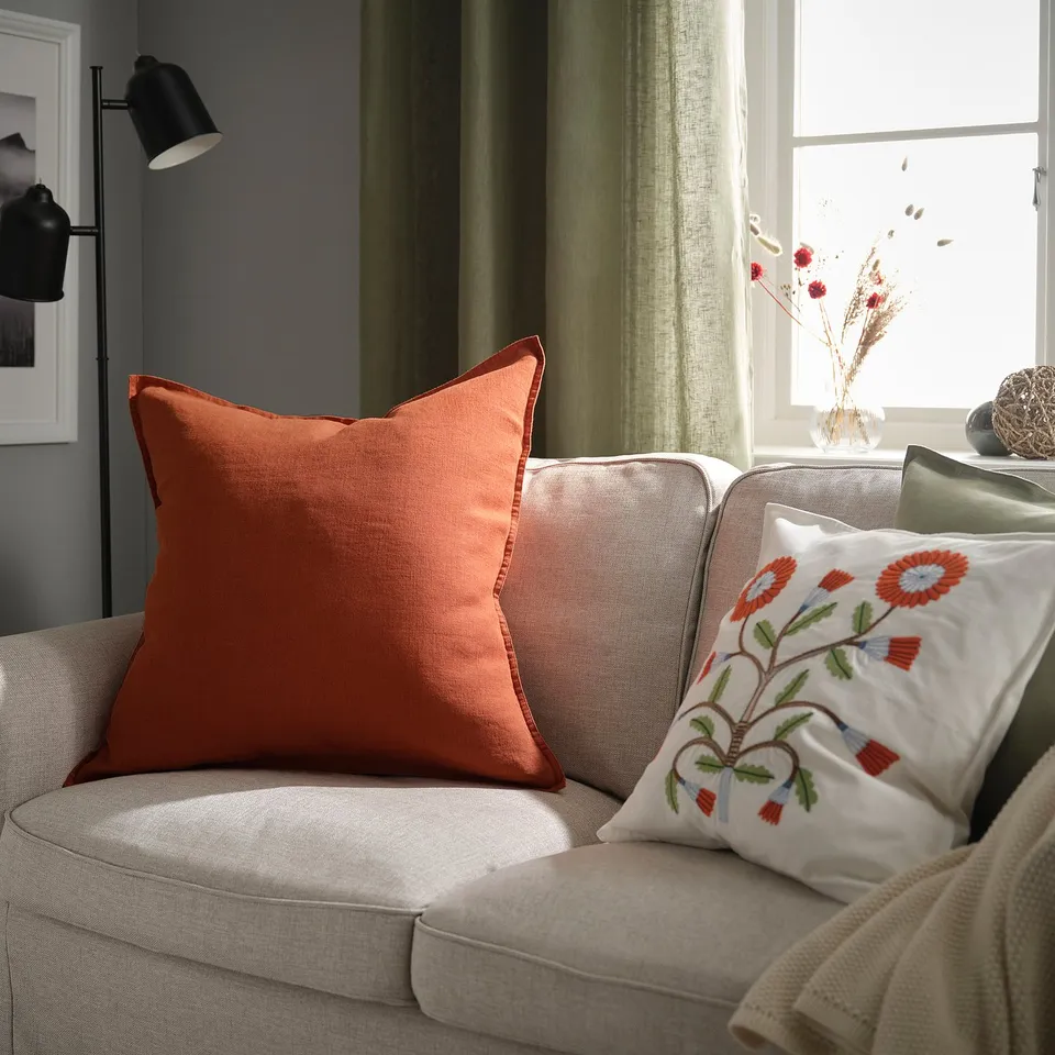 A cosy living room with a large red-brown linen cushion, embroidered floral pillow, and beige sofa. The curtain is green, and a lamp is on the left.