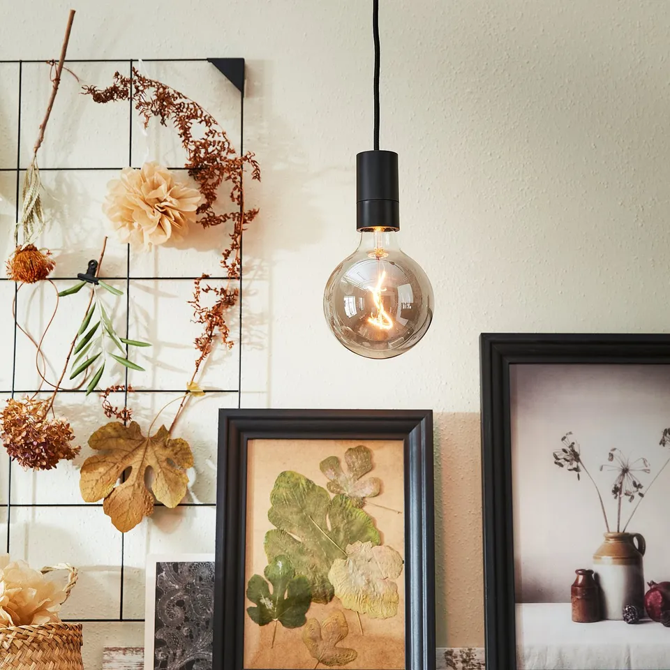 A cosy room with a hanging black SUNNEBY led bulb, emitting warm light through smoke-colored glass, surrounded by framed floral art and dried flowers.