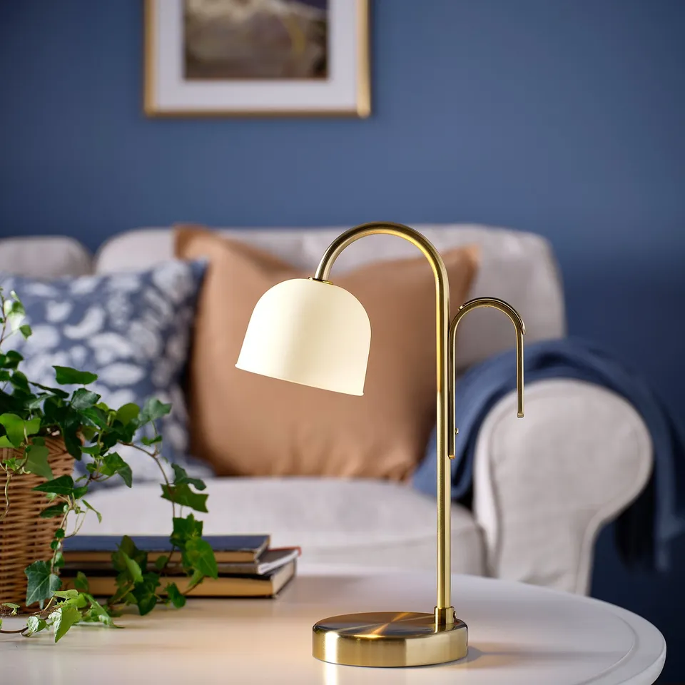 TVÅMASTAD lamp with brass base and frosted glass on table beside couch, near books and plant.