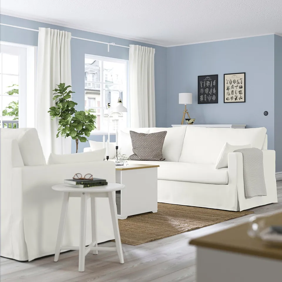 Modern living room with HYLTARP white sofa, pale blue walls, white curtains, side table with books, plant, and framed art.