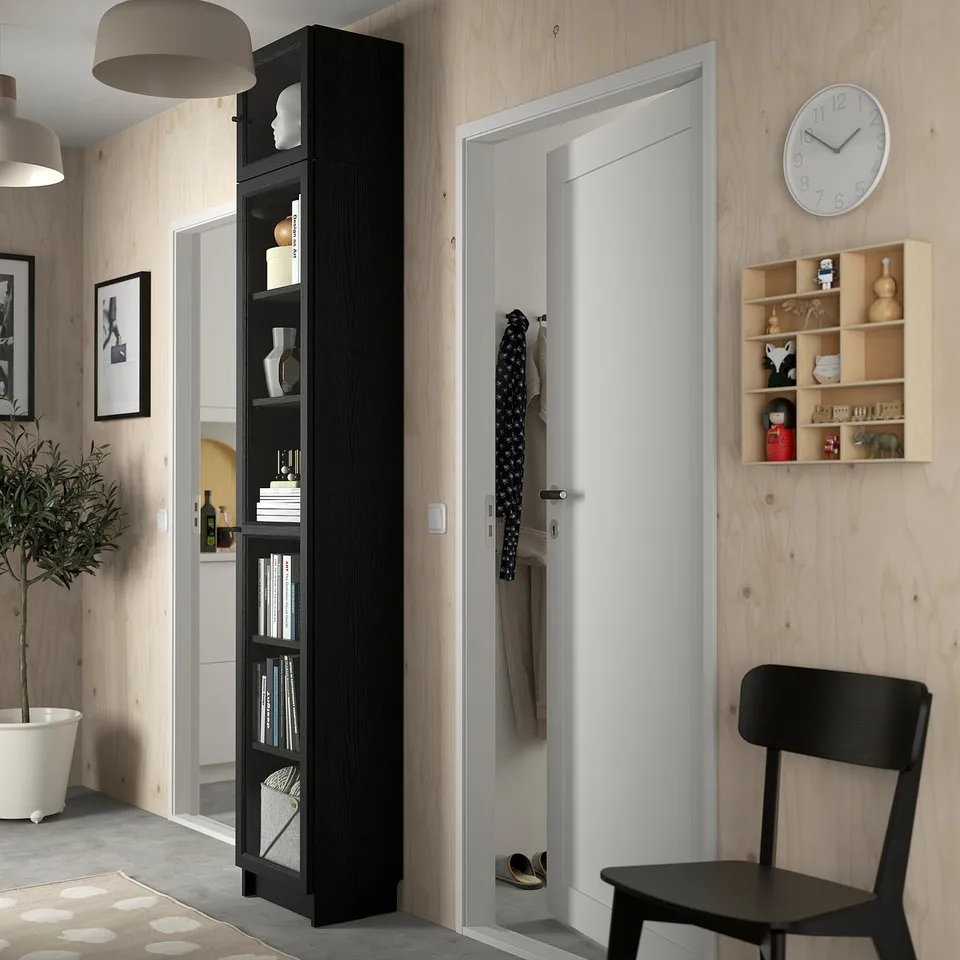 Minimalist entryway with tall black BILLY bookcase, white door, wall clock, black chair, and plant. Wooden walls.