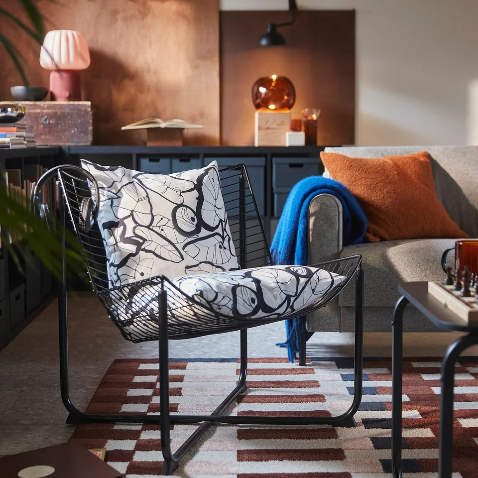 Black-white leaf-patterned cushion on metal chair; orange sofa, rug, decor in background.