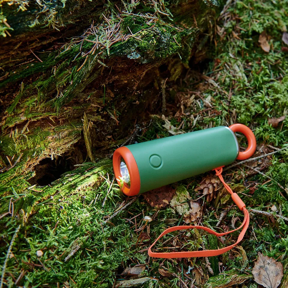 Green camping torch on mossy log, orange ring and strap, robust design.