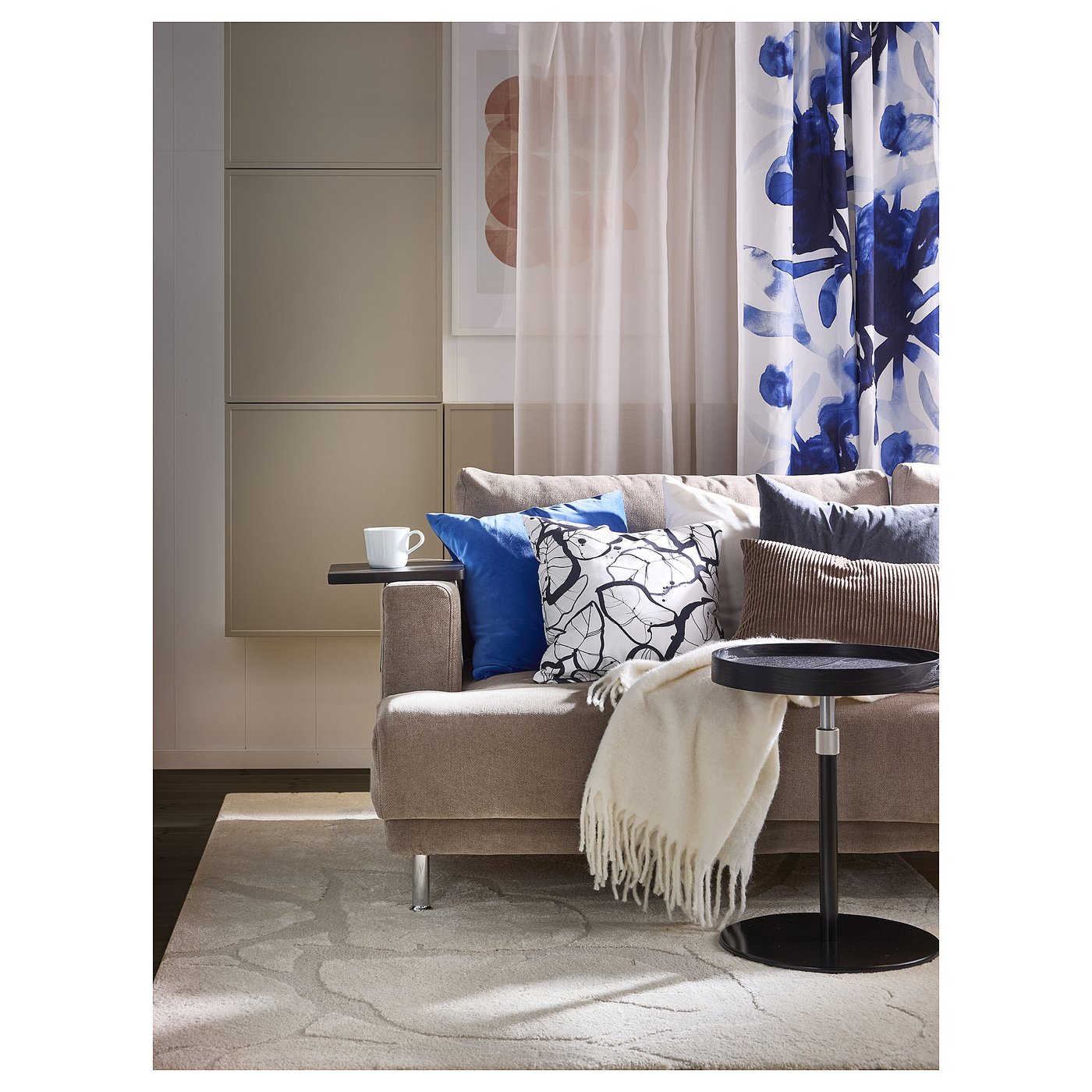 Modern living room with grey sofa, blue and white patterned cushions, black side table, white rug, and blue floral curtains.
