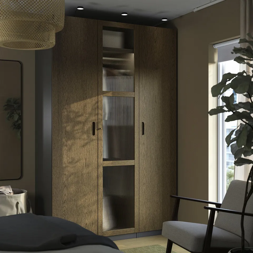 Modern bedroom with tall wooden PAX wardrobe, grey chair, plant, and window.