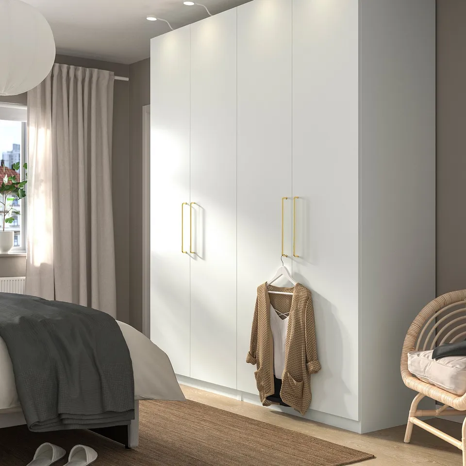 Modern bedroom with white PAX wardrobe, gold handles, sweater, dark/light linens bed, wicker chair near sheer curtains.
