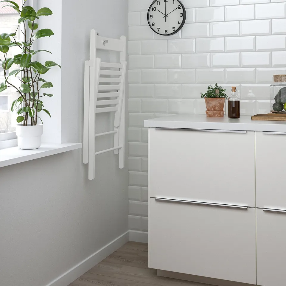 Minimalist kitchen with white cabinets, potted plant, and wall clock. Foldable white chair leans against wall.