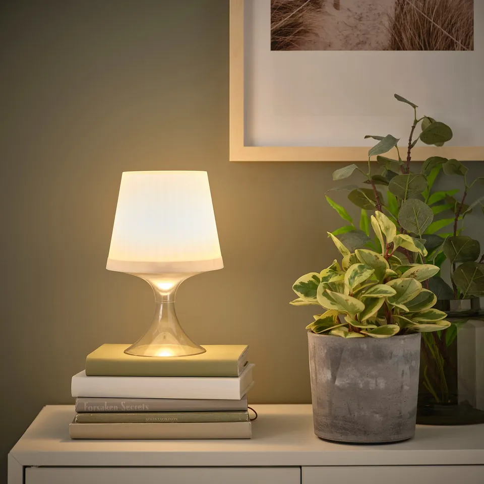 A modern table lamp sits atop a stack of books, casting warm light. Beside it, a potted plant adds greenery.