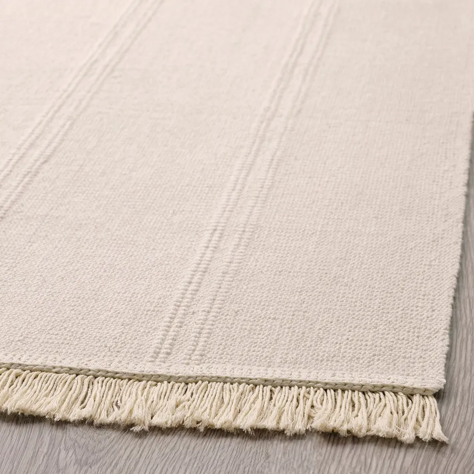 Beige cotton rug, neatly folded, light fringe edges, soft texture, flat weave.