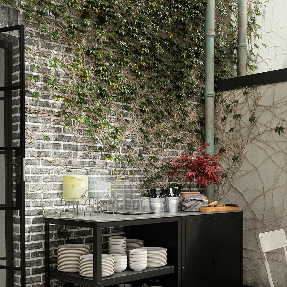 Black steel outdoor kitchen island with stainless steel top, storing white dishes, glasses, and lanterns. Hanging plants and brick wall background.