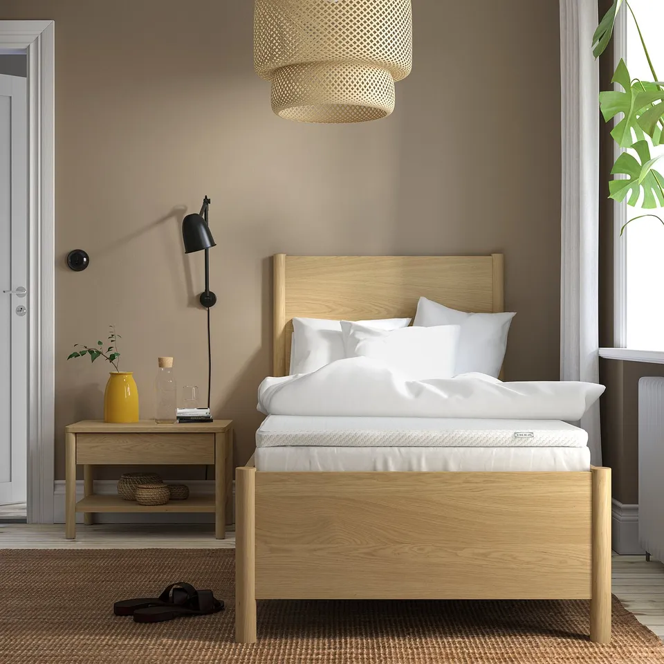 Minimalist bedroom: light wood bed, white linens, black lamp, yellow vase, rug, large window, brown walls.