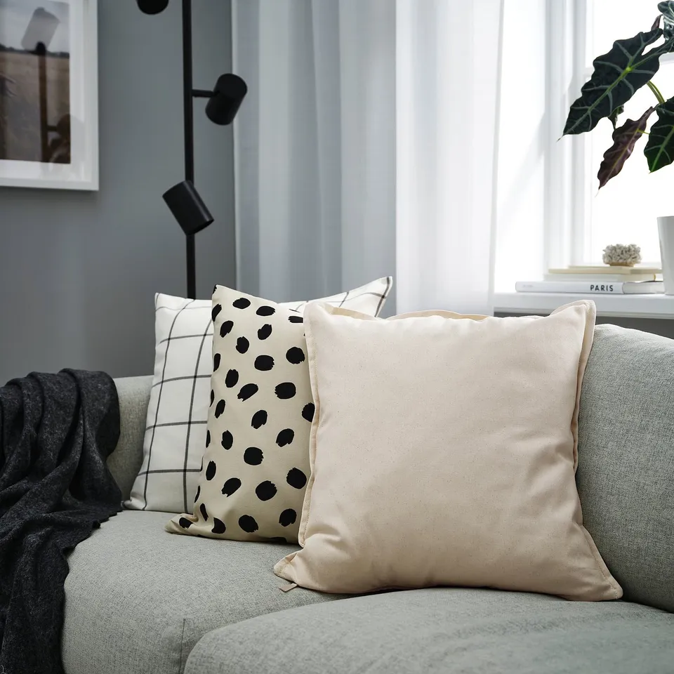 Modern living room with GURLI beige, black dot, and grid pillows on a grey couch near a window, plant, and black floor lamp.