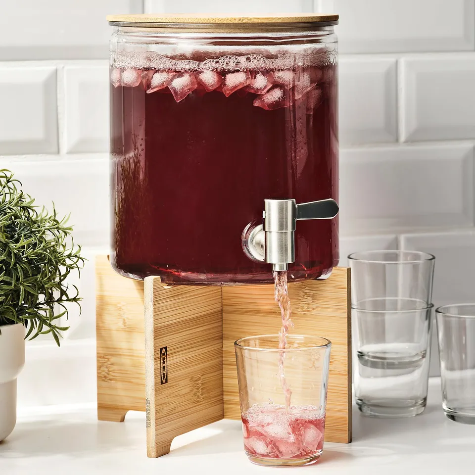 Glass jar with beverage dispenser and ice, pouring drink into glass on wooden stand.