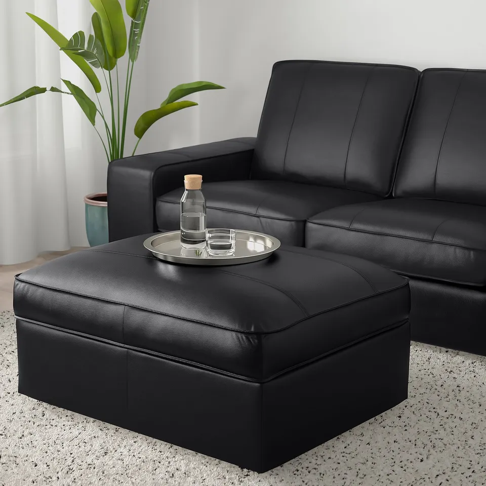 Black KIVIK leather footstool with storage, matching black sofa, clear tray with glass items on it, green plant beside, white curtains.