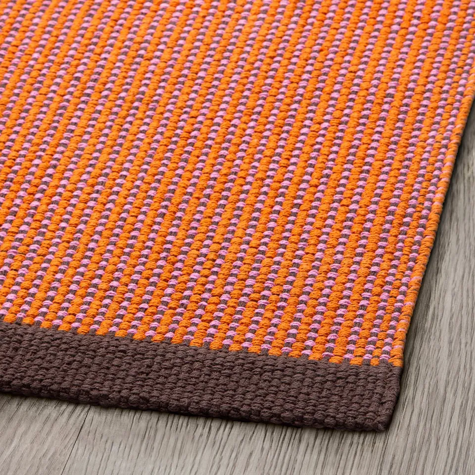 Grid-patterned rug with orange, pink, and brown colours laid on wooden floor. Dense weave, flat surface, and neat edges indicate high quality.