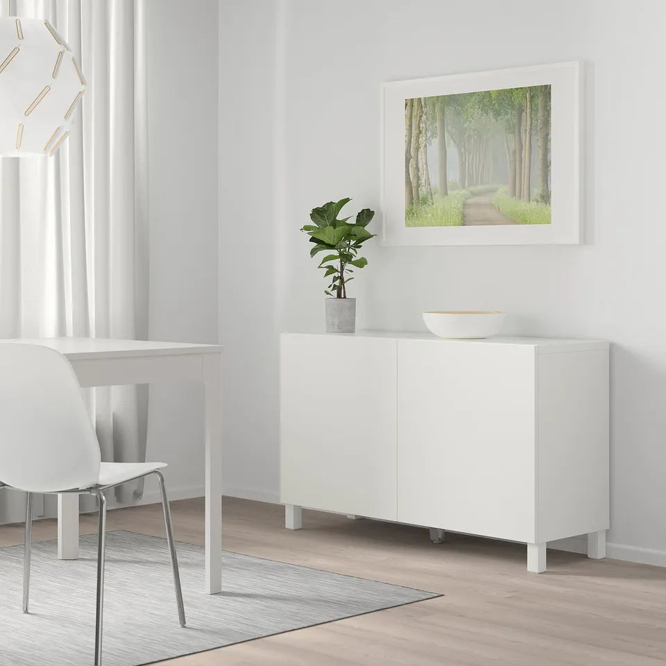 Modern minimalist dining room with white BESTÅ sideboard, table, chair, and rug. Wooden floor, plant, bowl, and forest art. Soft light.