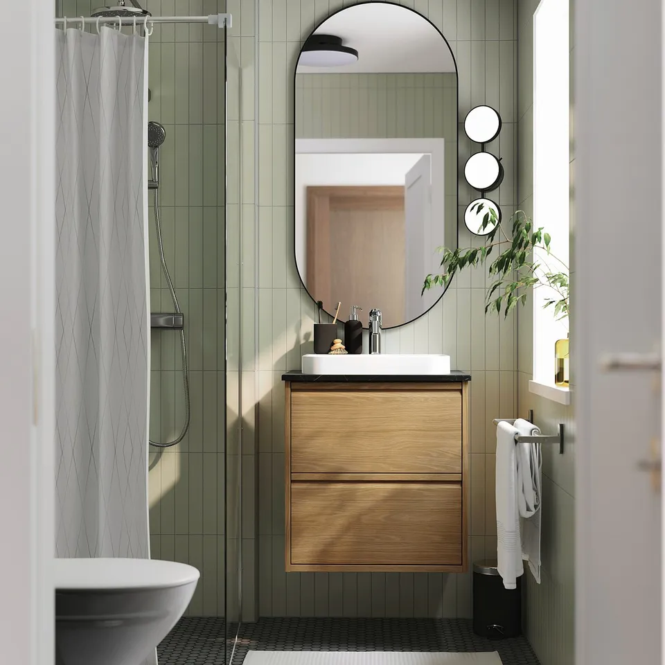 Modern bathroom with ÄNGSJÖN furniture: wooden cabinet, marble sink, glass shower, green tiles, plant, & mirrors.