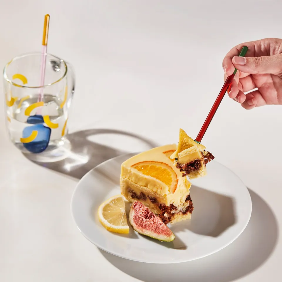 A hand lifts a dessert slice with FENGÖMMARE glass spoon. The spoons coloured design adds vibrancy. A plate with fruit slices accompanies.