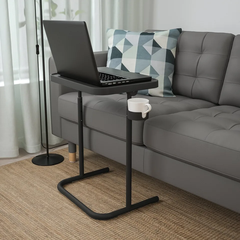 Black BJÖRKÅSEN laptop stand between grey couch pillows. Small, adjustable, with white cup holder.
