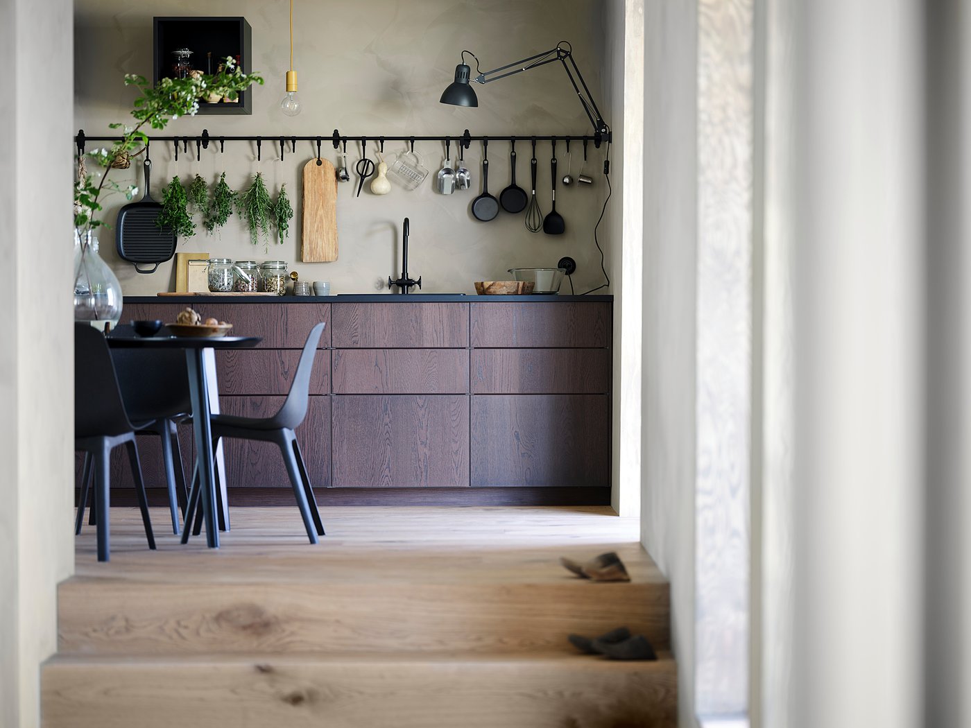 Modern kitchen with dark wooden cabinets, hanging utensils, black chairs, and a dining table.