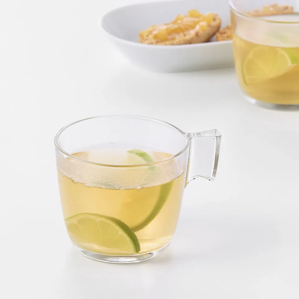 Clear glass mug with lime slices, holding a refreshing drink. Two cups on a white table with a snack bowl nearby.