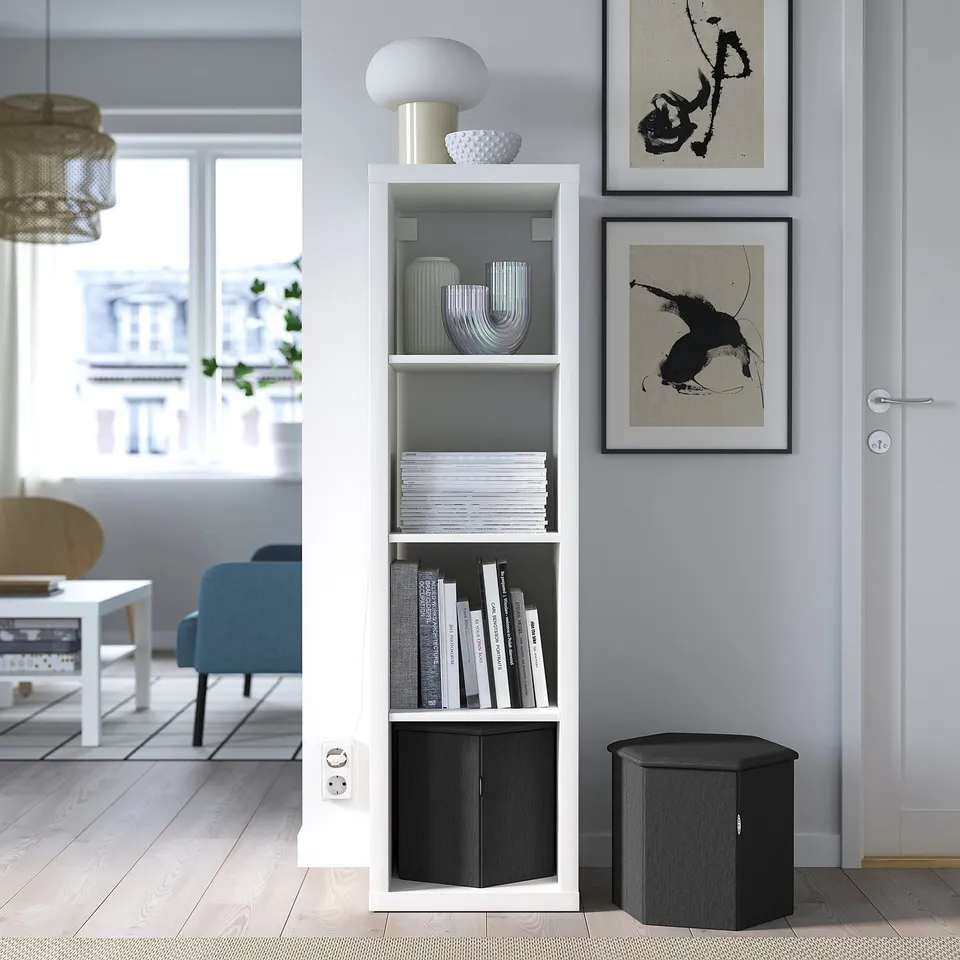 Modern living room with KJUGE ottoman, white bookshelf, blue couch, black & white art, large window, wooden floor.