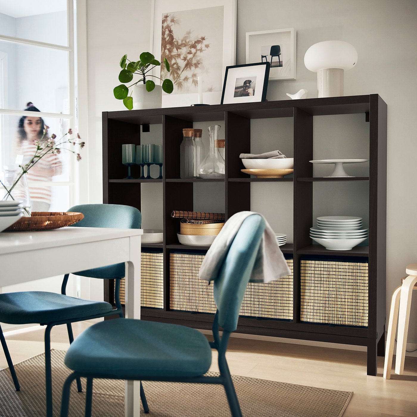 Modern dining area with KALLAX shelf, dishes, decor, blue chairs, person organising on white table. Bright, minimalist decor.