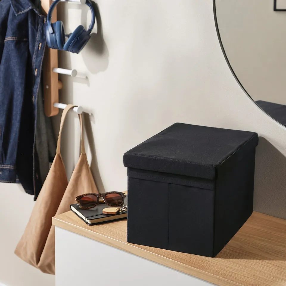 Black fabric storage box on a wooden shelf with sunglasses, phone, and a notebook on top. A tan handbag hangs beside it.