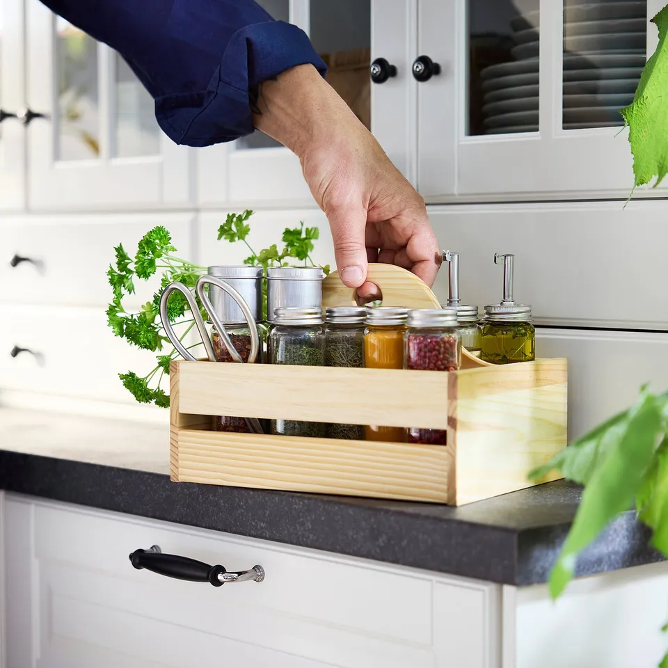 Hand fetching spice jar from wooden organiser on kitchen counter. Organiser holds various jars and fresh herbs.