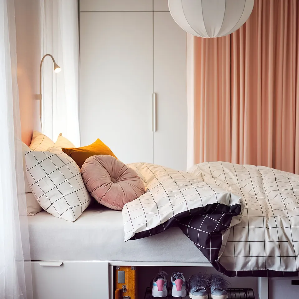 Cosy bedroom with light-colored bedding, colourful pillows, white pendant light, and yellow box beneath.