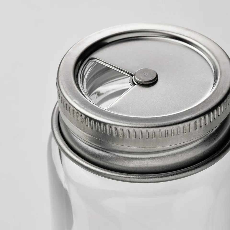 Close-up of a clear glass jar with a stainless steel lid, featuring a small metal button in the centre. The lid has a textured edge for easy grip.