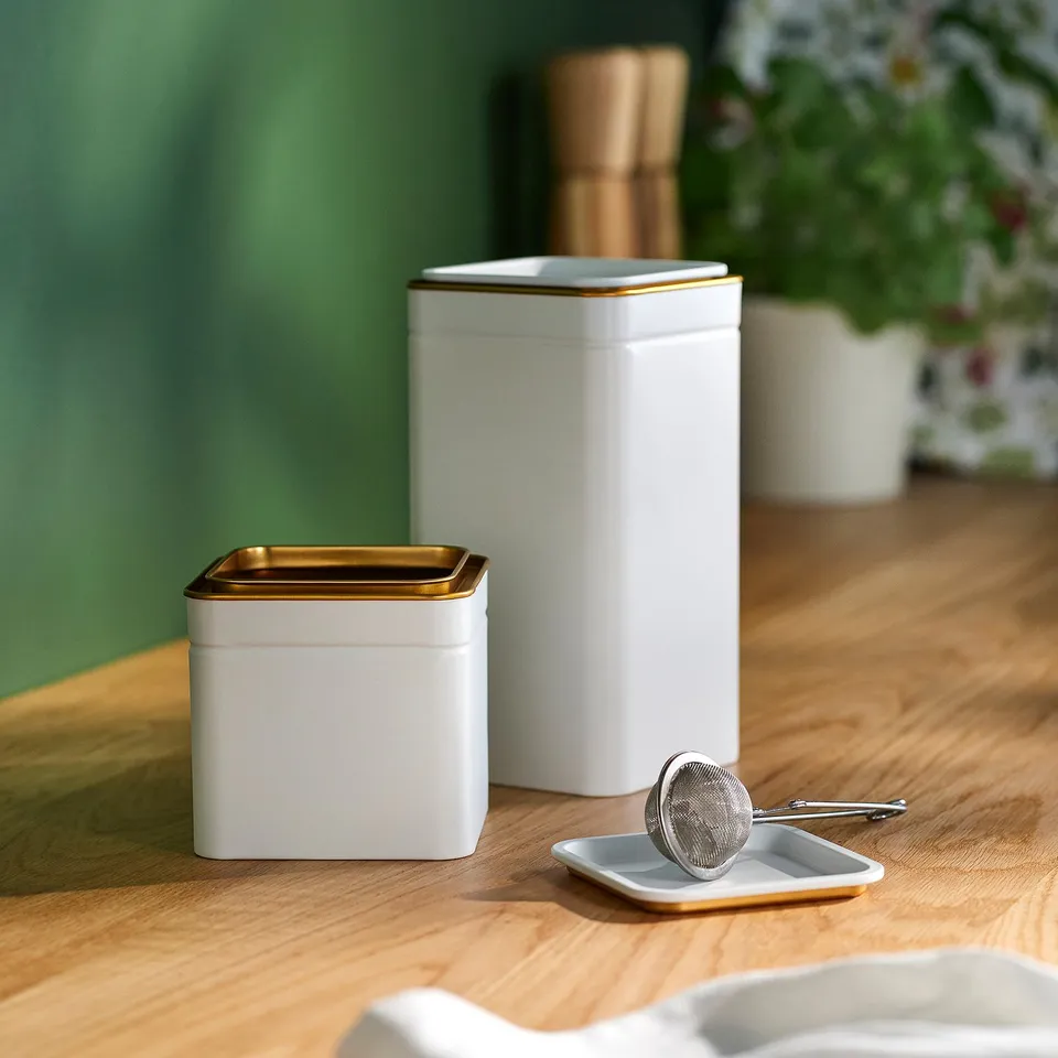 BLOMNING white metal tins: square, gold trim, stackable, shown on wooden table with a tea strainer, capable of storing tea, coffee, or spices.