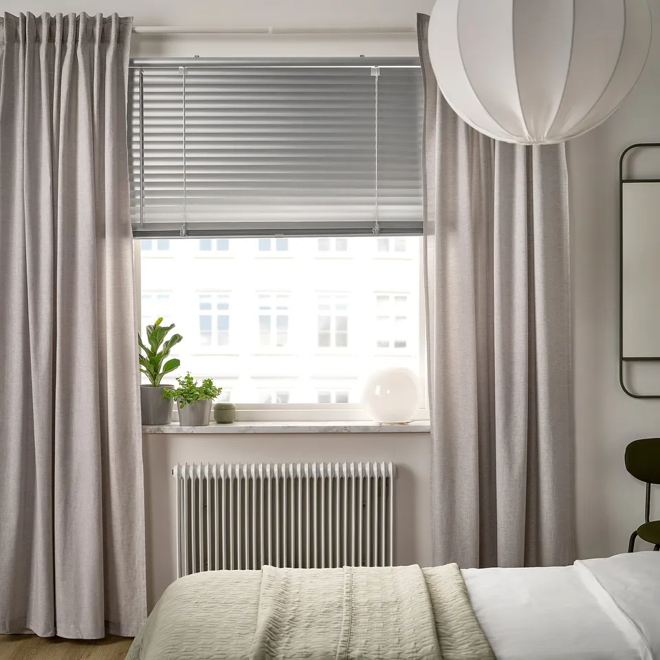 Modern bedroom with large window. Vecklary white blinds, light grey curtains, cream bed. Small potted plant on sill.