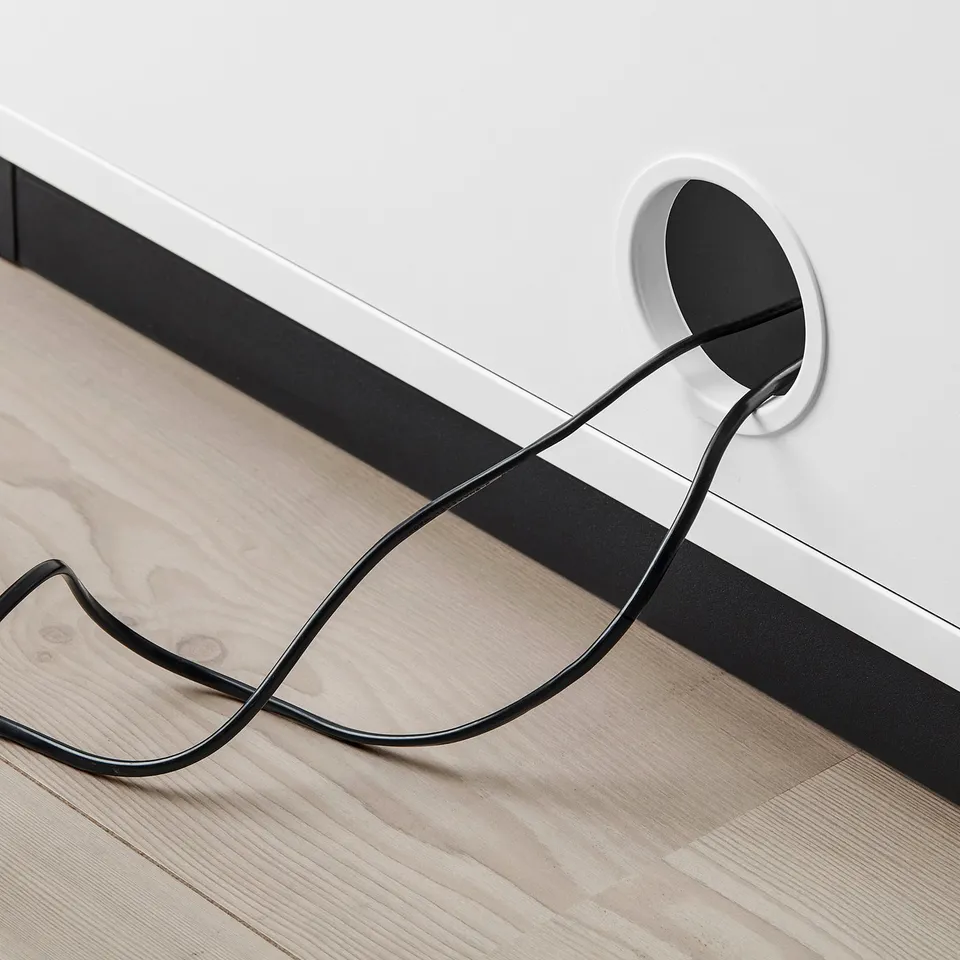 Black power cable exiting circular hole in white cabinet, lying on wooden floor.