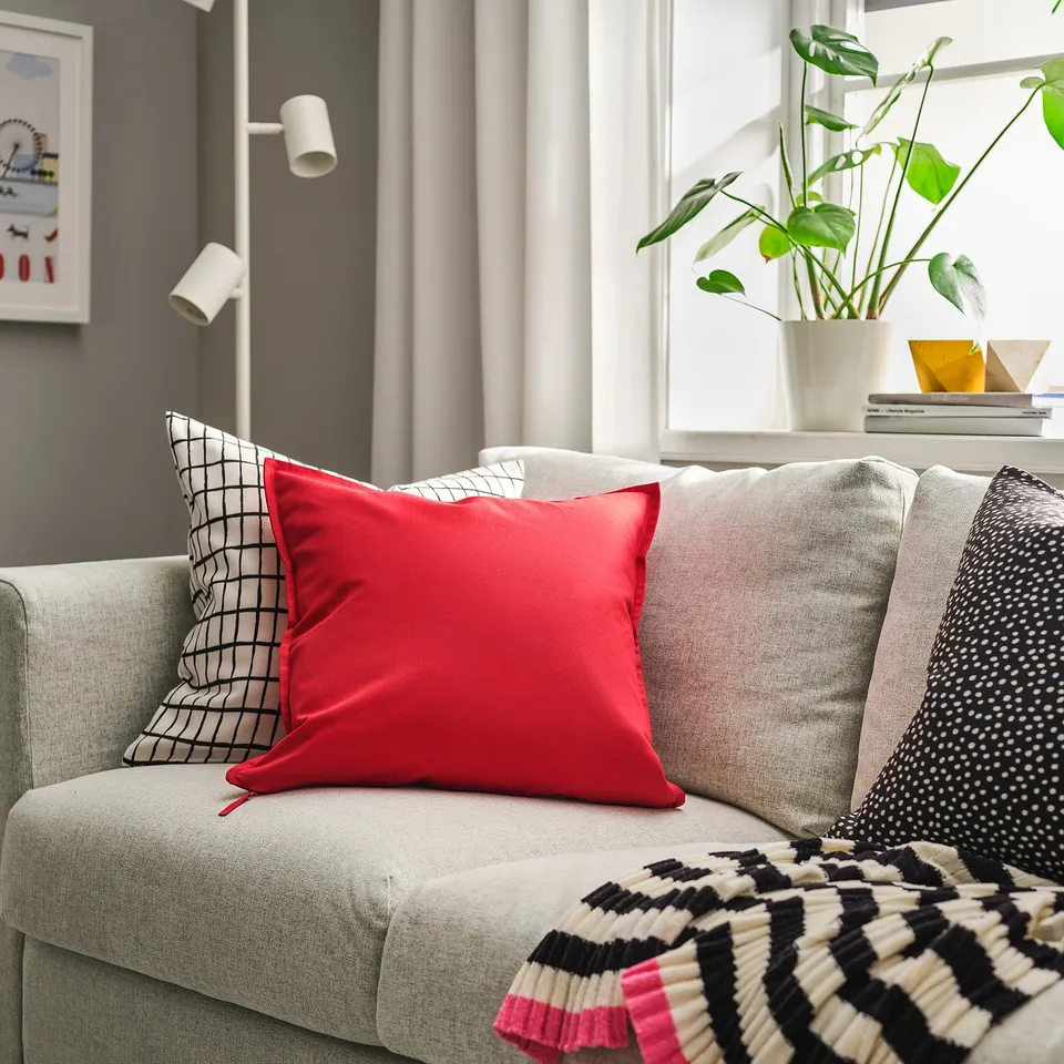 A cosy living room with a modern couch, a red GURLI pillow, striped blanket, plant, and books.
